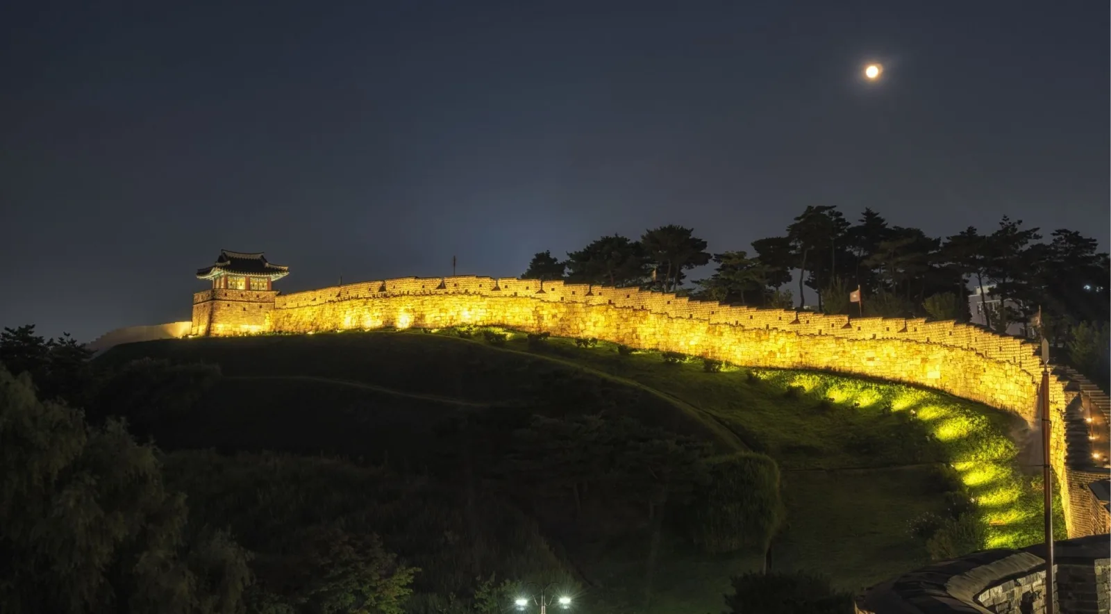 The Hwaseong Fortress wall floodlit at night with the moon above