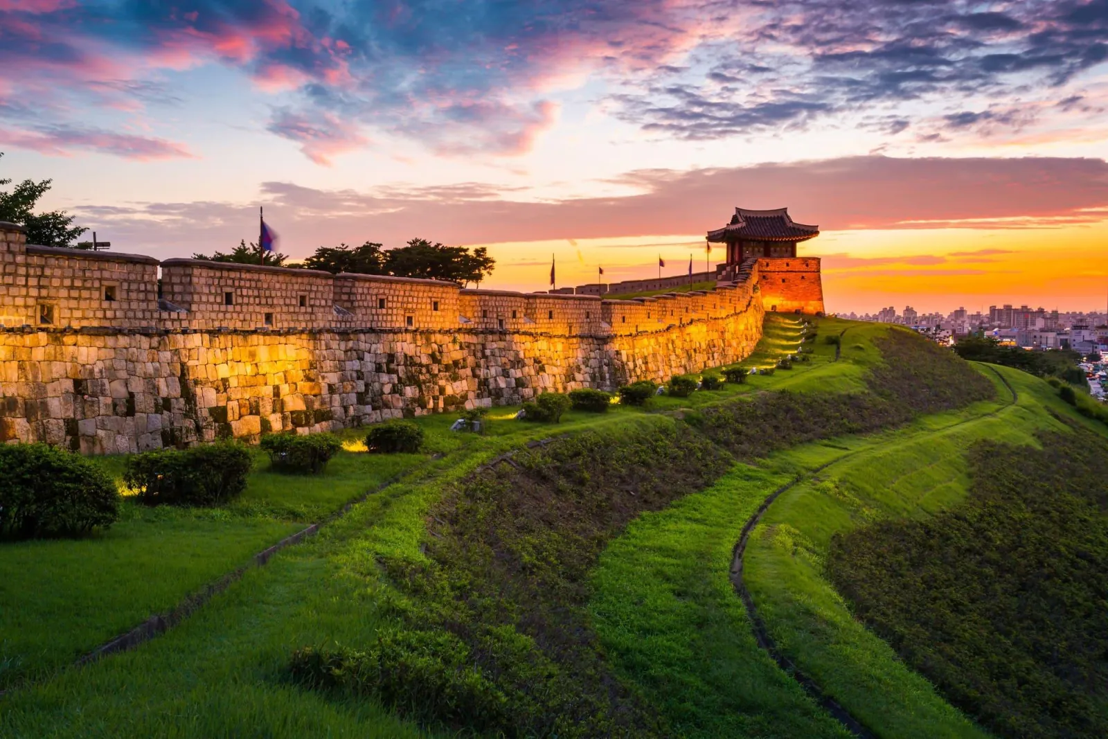 Floodlit fortress rampart and grass embankment at Hwaseong under a pink sky